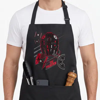 Pilot tie apron - Cartoon stormtrooper helmet with red TIE fighter text and imperial emblem in black and white — shown on a black cotton canvas apron against a white background