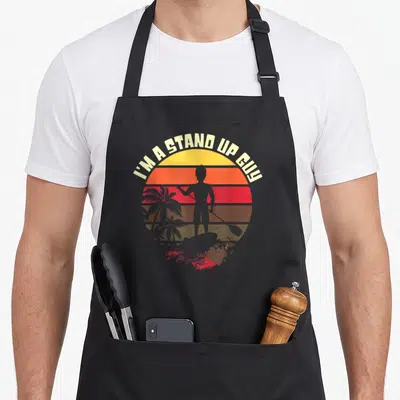 Board stand up paddle apron - Retro sun with man standing on surfboard, palm trees, text I'M A STAND UP GUY in cream — shown on a black cotton canvas apron against a white background