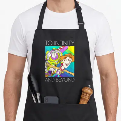 Woody infinity apron - Buzz Lightyear and Woody cartoon flying against rainbow stripes with TO INFINITY AND BEYOND text — shown on a black cotton canvas apron against a white background