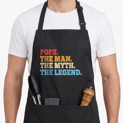 Father pop grandpa apron - Stacked text POPS THE MAN THE MYTH THE LEGEND in red yellow beige and blue — shown on a black cotton canvas apron against a white background