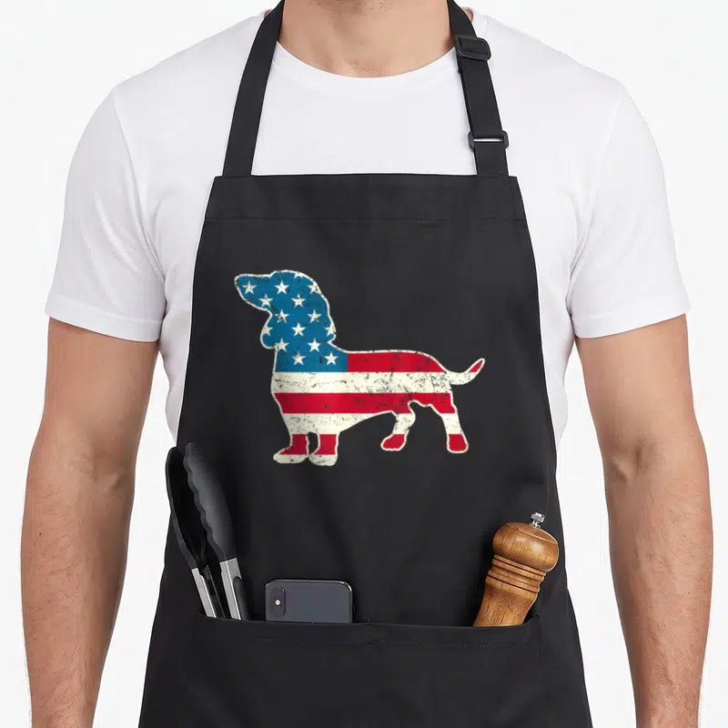 Dachshund 4th of july apron - Distressed American flag pattern filling a standing dachshund silhouette in blue red — shown on a black cotton canvas apron against a white background
