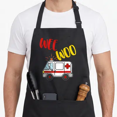 Ems ambulance girl apron - Cartoon white ambulance with red cross and arched WEE WOO text in red and yellow — shown on a black cotton canvas apron against a white background