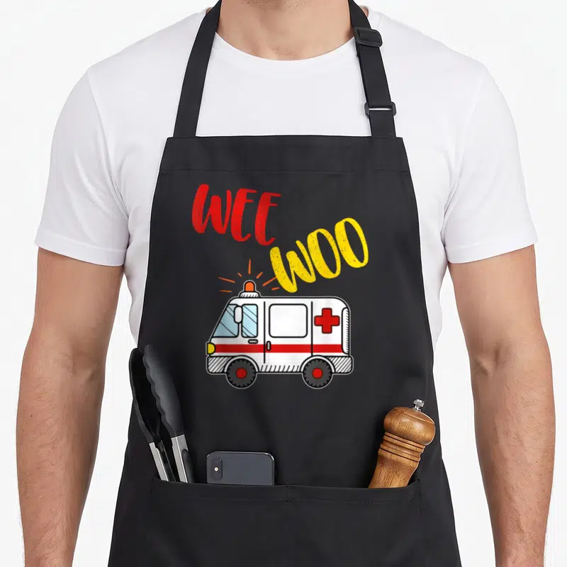 Ems ambulance girl apron - Cartoon white ambulance with red cross and arched WEE WOO text in red and yellow — shown on a black cotton canvas apron against a white background