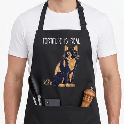 Tortoise cat apron - Sitting tortoiseshell cat with white, black, and orange fur below white text reading TORTITUDE IS — shown on a black cotton canvas apron against a white background