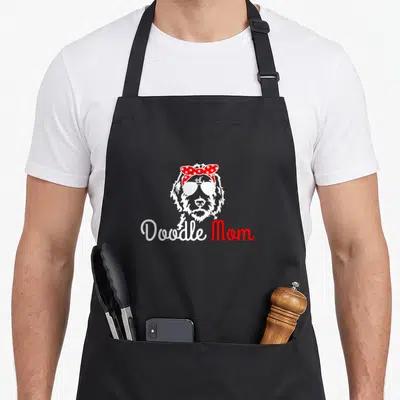 Golden dog apron - Cartoon doodle face with sunglasses and red polka dot bow above Doodle Mom text — shown on a black cotton canvas apron against a white background