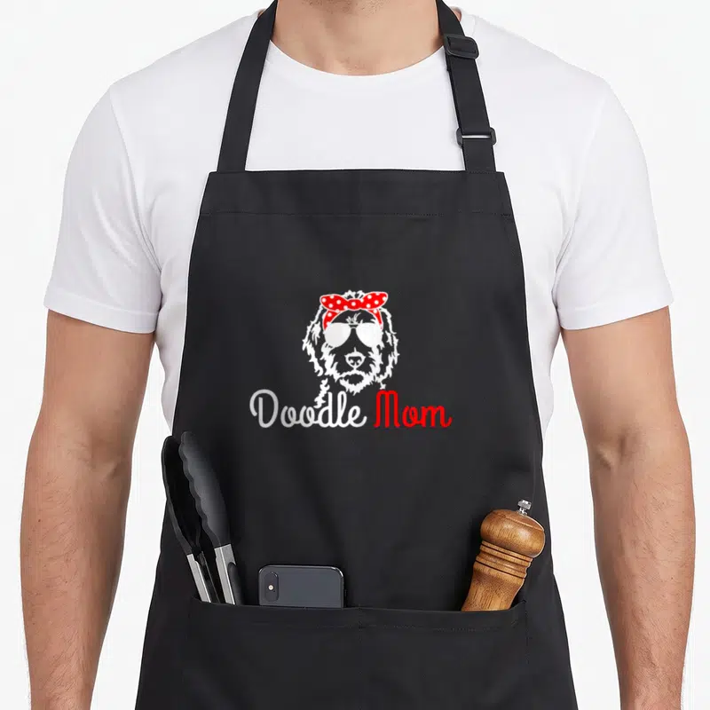 Golden dog apron - Cartoon doodle face with sunglasses and red polka dot bow above Doodle Mom text — shown on a black cotton canvas apron against a white background