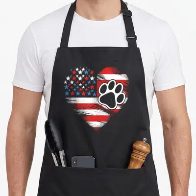 Paw 4th of july apron - Distressed heart-shaped USA flag pattern with overlapping paw print in red white blue and grey — shown on a black cotton canvas apron against a white background