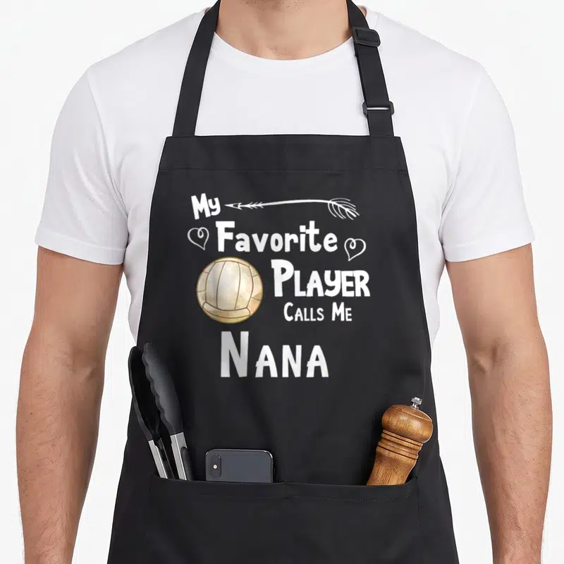 Volleyball nana aprons - Stacked white text My Favorite Player Calls Me Nana with beige volleyball and hearts — shown on a black cotton canvas apron against a white background