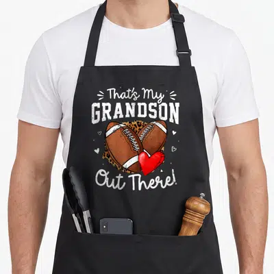 Grandson grandma aprons - That's My GRANDSON Out There text with crossed footballs and leopard print heart — shown on a black cotton canvas apron against a white background
