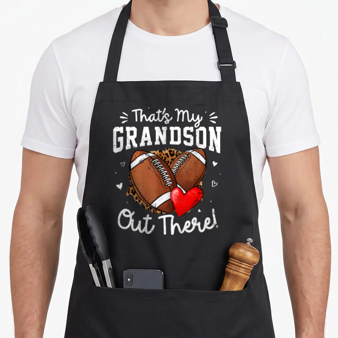 Grandson grandma aprons - That's My GRANDSON Out There text with crossed footballs and leopard print heart — shown on a black cotton canvas apron against a white background