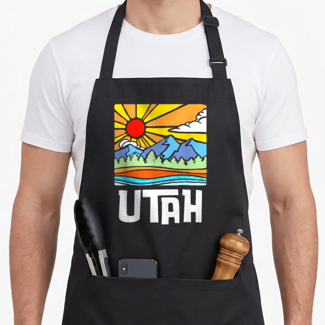Utah artistic apron - Retro square graphic of sun, blue mountains, green trees, and water above white UTAH text — shown on a black cotton canvas apron against a white background