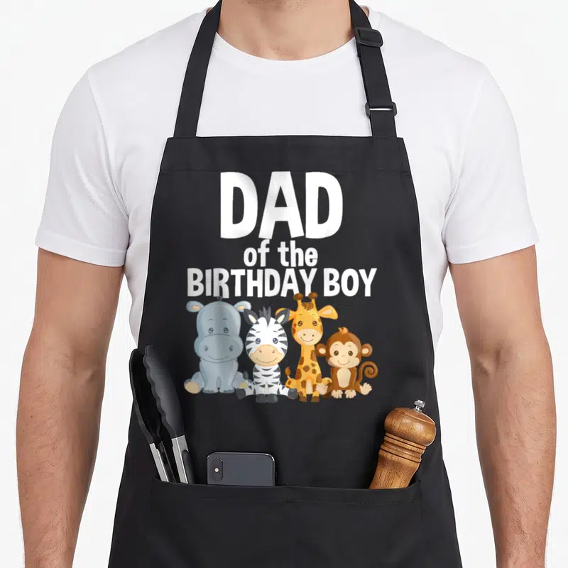 Jungle safari birthday apron - Dad of the Birthday Boy text above cartoon hippo zebra giraffe and monkey — shown on a black cotton canvas apron against a white background