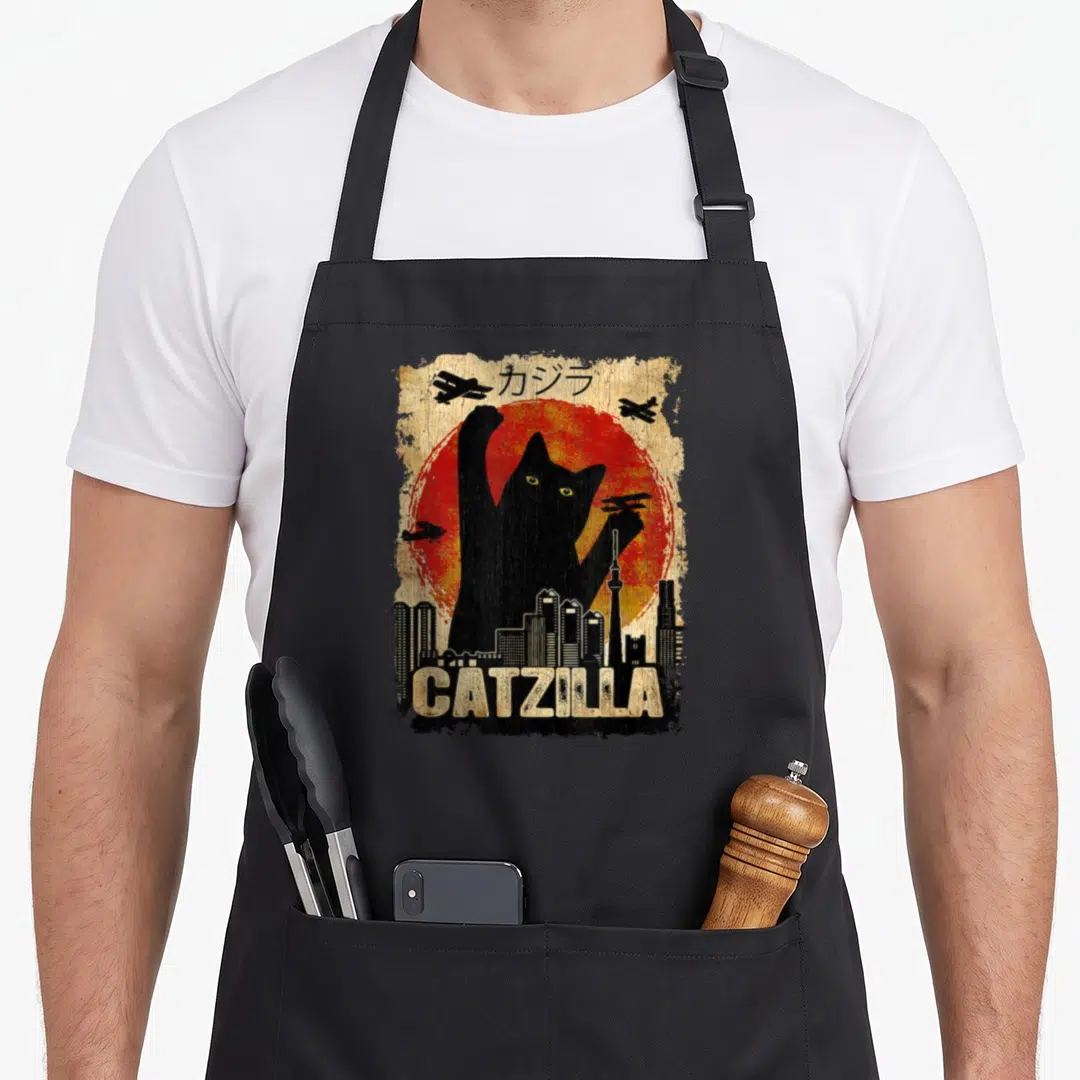Catzilla kitten apron - Giant black cat silhouette against red sun with CATZILLA text and skyline — shown on a black cotton canvas apron against a white background