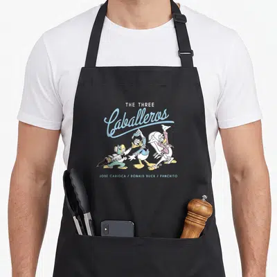 Three duck apron - The Three Caballeros cartoon print with blue script and three standing ducks labeled by name — shown on a black cotton canvas apron against a white background