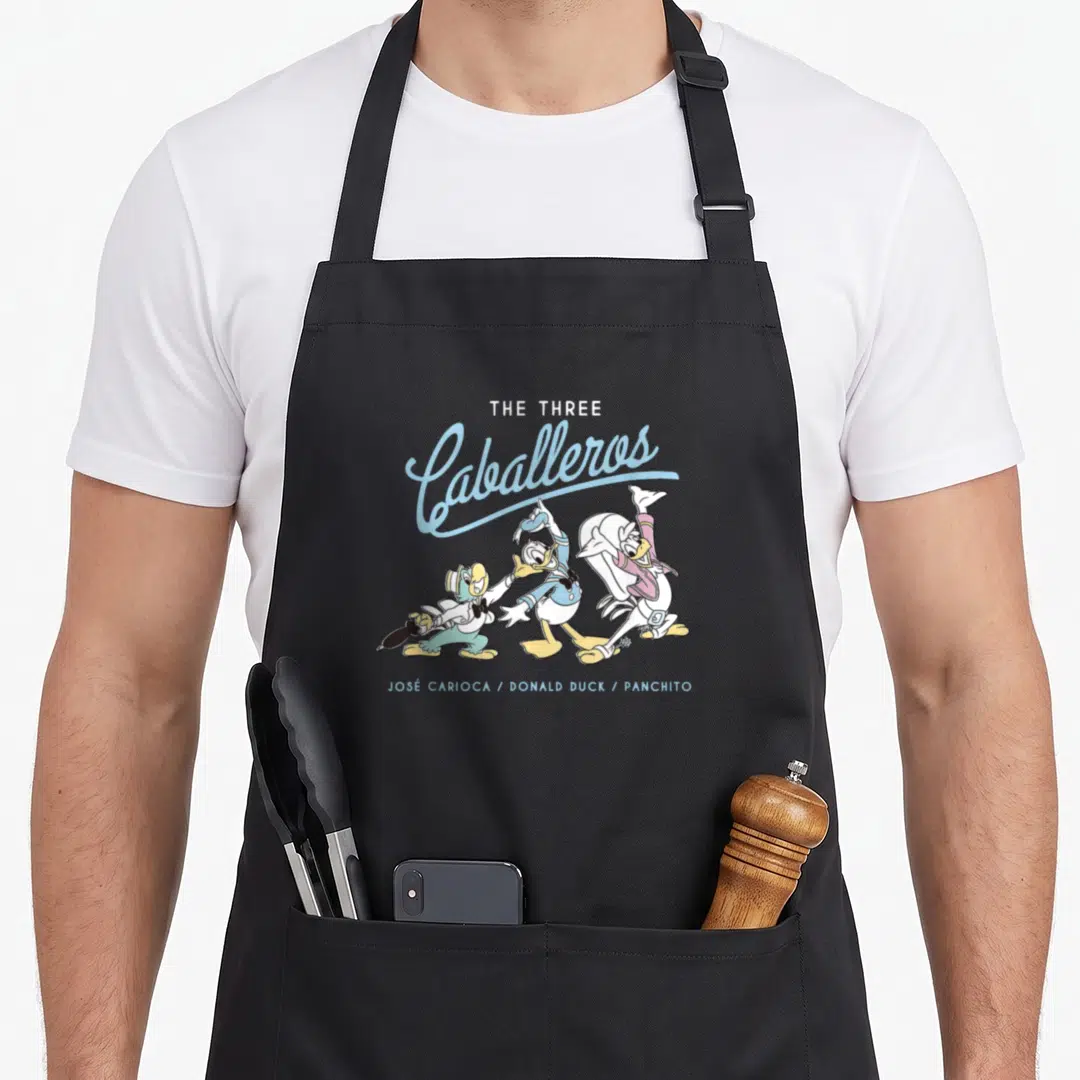 Three duck apron - The Three Caballeros cartoon print with blue script and three standing ducks labeled by name — shown on a black cotton canvas apron against a white background