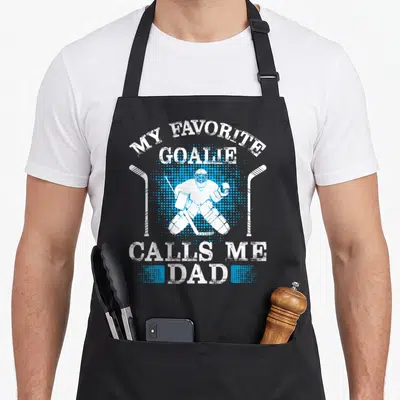 Goalie winter apron - Distressed white text My Favorite Goalie Calls Me Dad surrounding goalie silhouette and two — shown on a black cotton canvas apron against a white background
