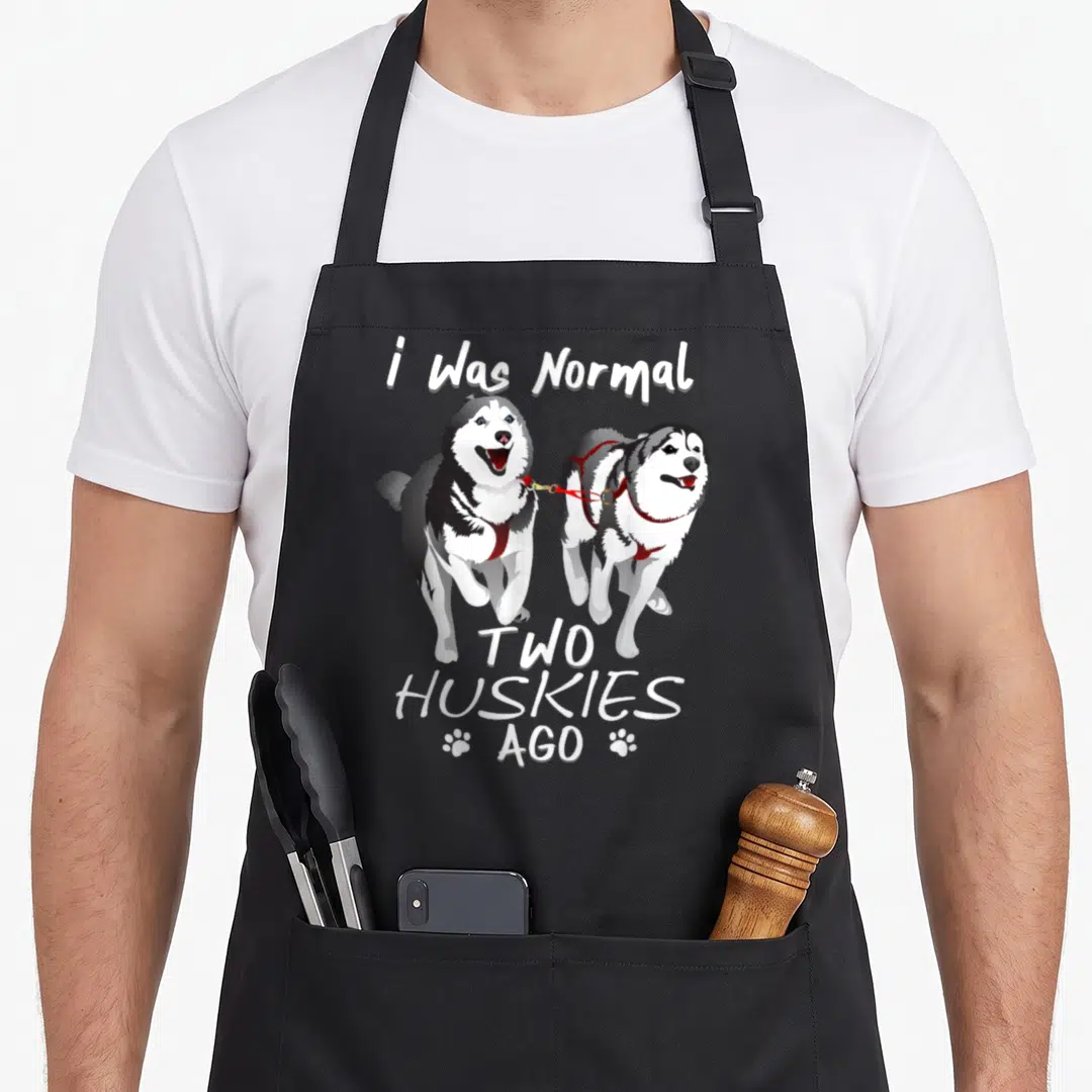 Normal husky apron - Two grey and white huskies on red leash with i Was Normal TWO HUSKIES AGO text — shown on a black cotton canvas apron against a white background