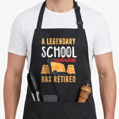 School retirement apron - Cartoon floor scrubber between caution signs with text A LEGENDARY SCHOOL custodian HAS — shown on a black cotton canvas apron against a white background