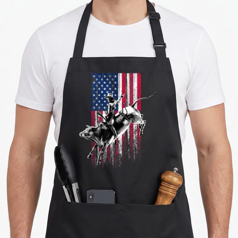 Rodeo american flag apron - Bull rider on bucking bull over distressed American flag in red white blue black — shown on a black cotton canvas apron against a white background