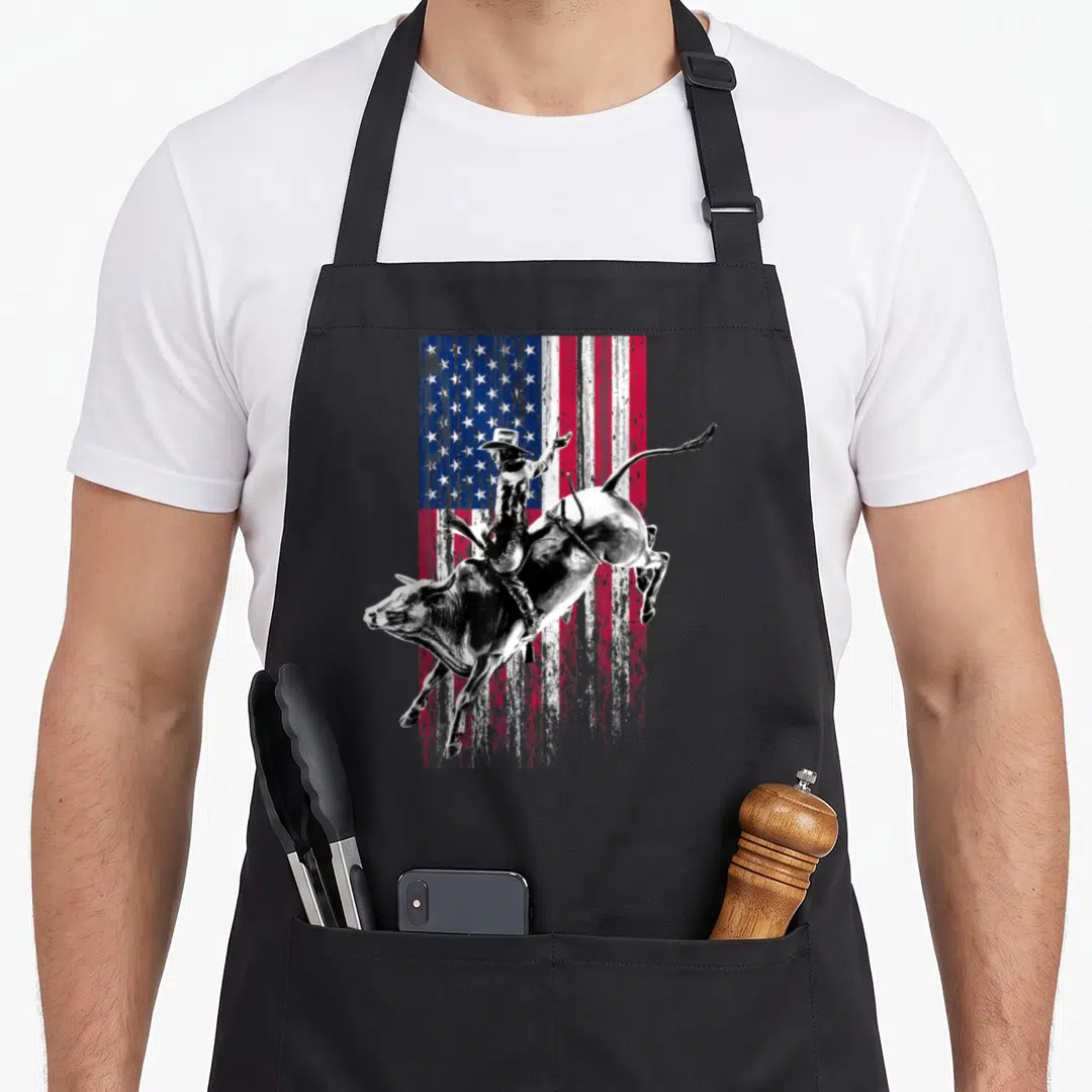 Rodeo american flag apron - Bull rider on bucking bull over distressed American flag in red white blue black — shown on a black cotton canvas apron against a white background