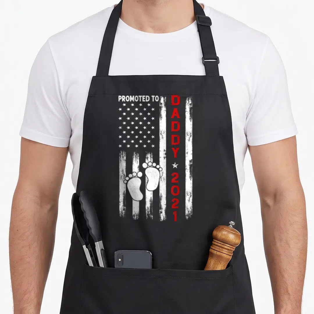 Promoted daddy father day apron - Distressed USA flag with baby footprints and Promoted To Daddy 2021 text — shown on a black cotton canvas apron against a white background