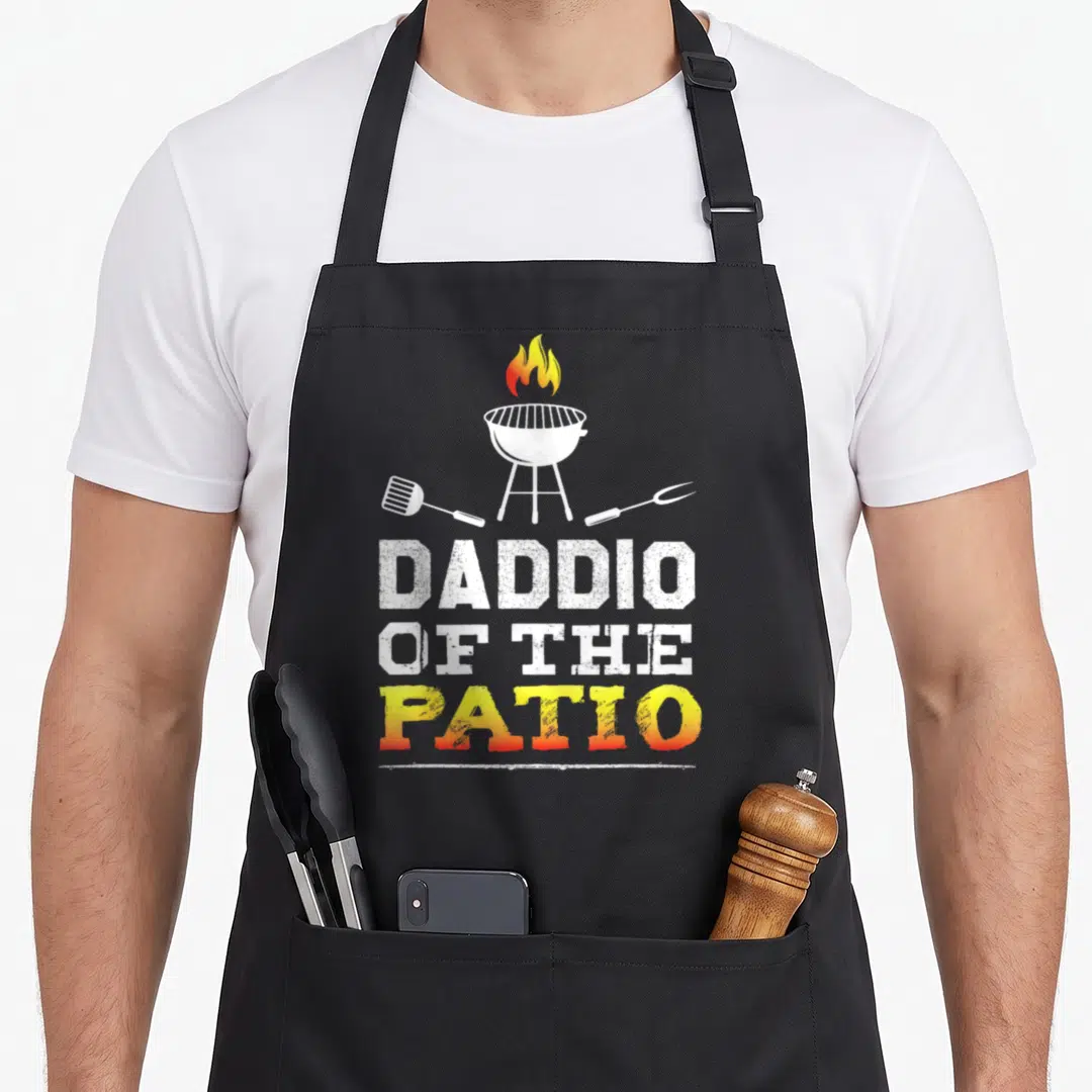 Daddio barbecue apron - Cartoon grill with flames, spatula, fork, and stacked text DADDIO OF THE PATIO — shown on a black cotton canvas apron against a white background