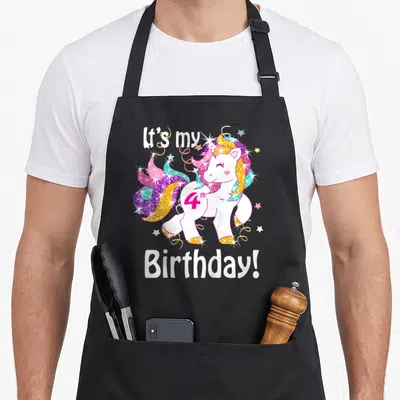 4th unicorn apron - White cartoon unicorn with pink purple blue yellow mane and 4th Birthday text surrounded by stars — shown on a black cotton canvas apron against a white background