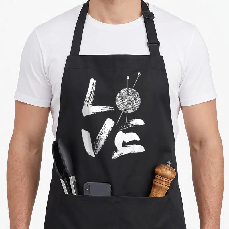 Needle knitting apron - White distressed LOVE text with yarn ball and two needles replacing the letter O — shown on a black cotton canvas apron against a white background