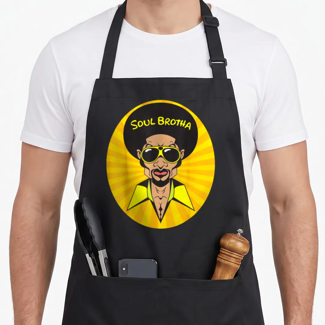Afro funky apron - Cartoon man with afro and sunglasses under SOUL BROTHA text on yellow sunburst — shown on a black cotton canvas apron against a white background