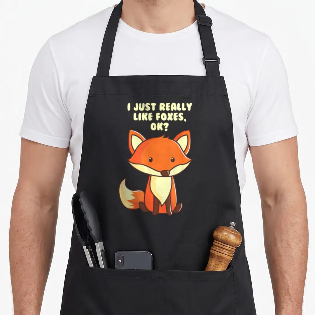 Funny fox aprons - Cartoon orange fox sitting below stacked text I Just Really Like Foxes Ok in pale yellow — shown on a black cotton canvas apron against a white background