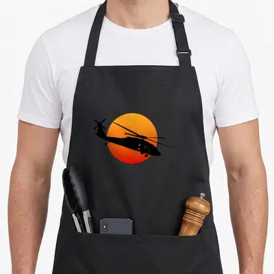 Hawk military apron - Black helicopter silhouette flying across an orange and red gradient sun circle — shown on a black cotton canvas apron against a white background
