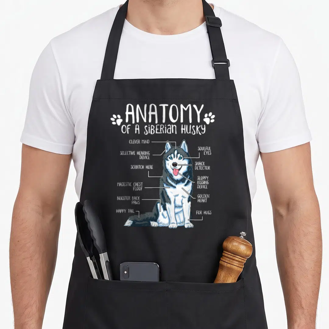 Anatomy husky apron - Cartoon diagram of a husky with labels for soulful eyes, snack detector, and golden heart — shown on a black cotton canvas apron against a white background