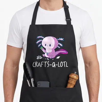 Lotl craft apron - Pink axolotl holding scissors and paper scraps with CRAFTS-a-LOTL text below — shown on a black cotton canvas apron against a white background