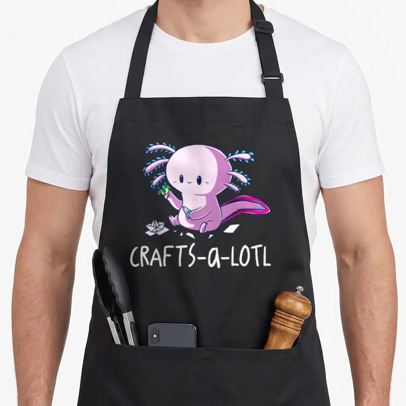 Lotl craft apron - Pink axolotl holding scissors and paper scraps with CRAFTS-a-LOTL text below — shown on a black cotton canvas apron against a white background