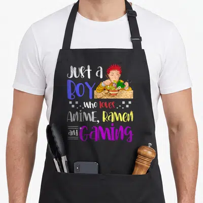 Gaming anime apron - Cartoon boy with red hair eating ramen and holding a game controller surrounded by colorful — shown on a black cotton canvas apron against a white background
