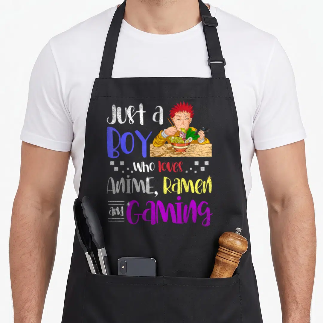 Gaming anime apron - Cartoon boy with red hair eating ramen and holding a game controller surrounded by colorful — shown on a black cotton canvas apron against a white background