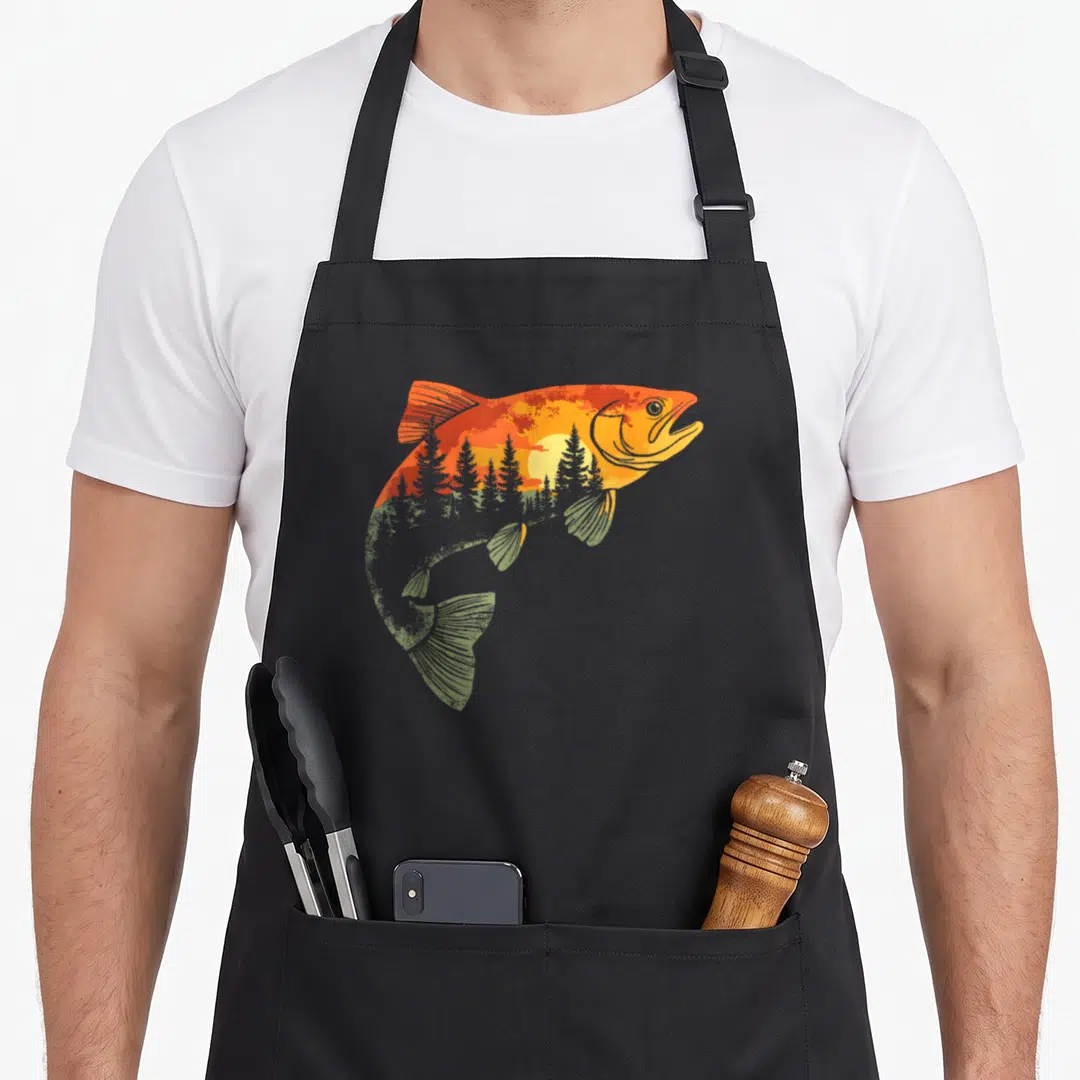 Nature fishing aprons - Fish silhouette filled with sunset, sun, and pine trees in orange and green — shown on a black cotton canvas apron against a white background