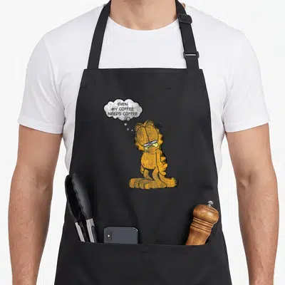 Coffee cat apron - Sleepy orange cartoon cat with thought bubble saying EVEN MY COFFEE NEEDS COFFEE — shown on a black cotton canvas apron against a white background