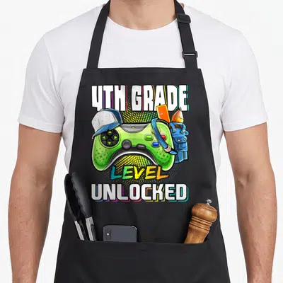 4th video game apron - Green game controller with blue backpack, orange ruler, pencil, and 4th Grade Level Unlocked — shown on a black cotton canvas apron against a white background