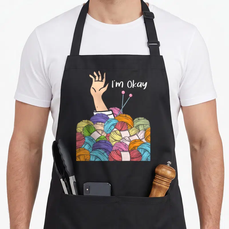 Okay knitting apron - Hand reaching from yarn pile with text I'm Okay in cartoon style — shown on a black cotton canvas apron against a white background
