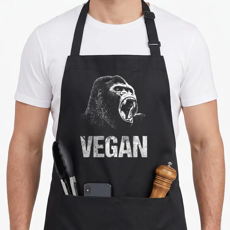 Food vegan apron - White distressed gorilla head roaring above VEGAN text on dark background — shown on a black cotton canvas apron against a white background