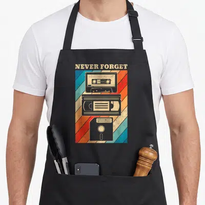 Floppy 80s apron - NEVER FORGET text above stacked cassette, VHS, and floppy disk on diagonal stripes — shown on a black cotton canvas apron against a white background