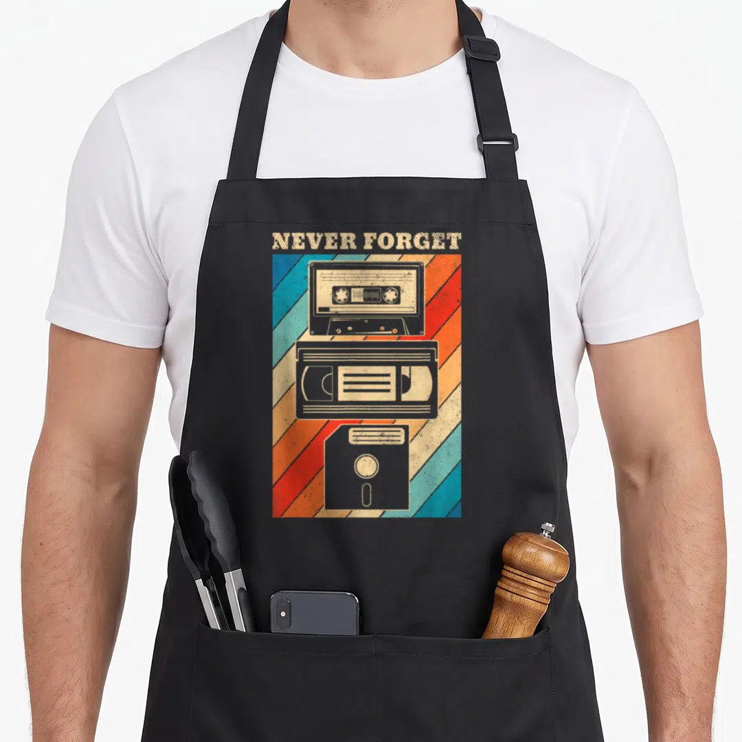 Floppy 80s apron - NEVER FORGET text above stacked cassette, VHS, and floppy disk on diagonal stripes — shown on a black cotton canvas apron against a white background