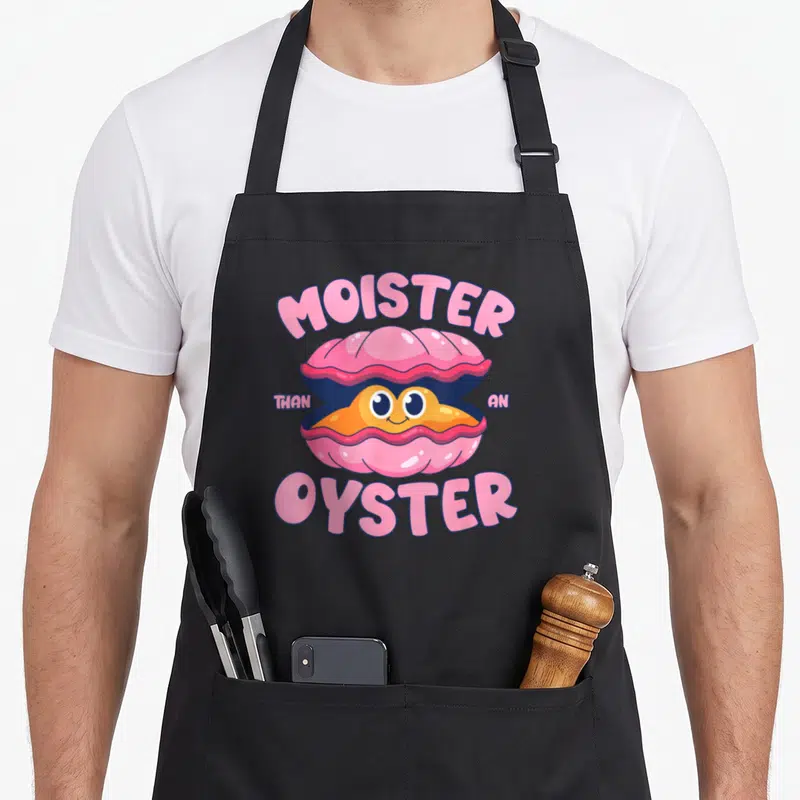 Bivalve oyster apron - Cartoon pink oyster with smiling face, orange meat, arched text reading MOISTER THAN AN OYSTER — shown on a black cotton canvas apron against a white background