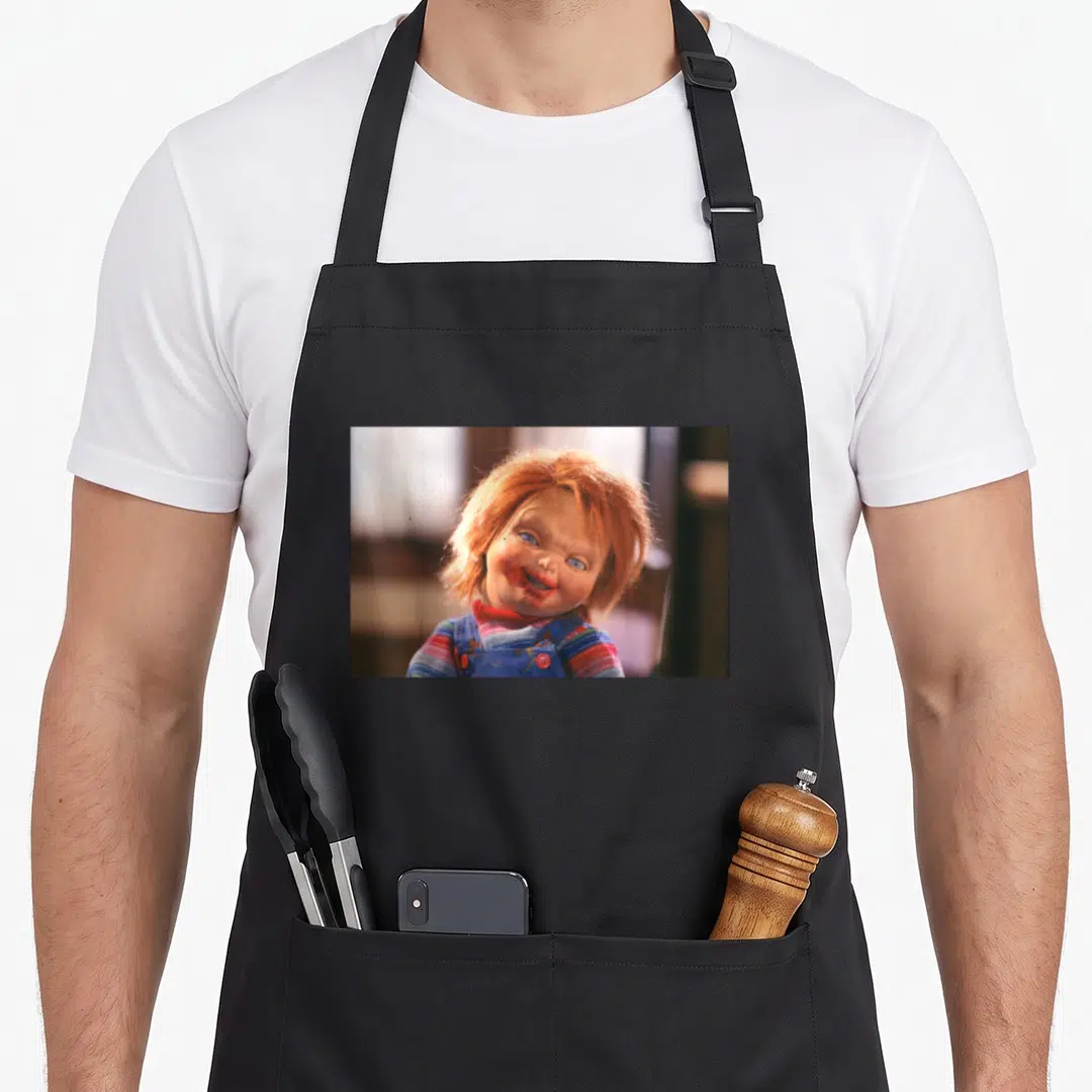 Face bloody apron - Photo print of doll with orange hair, blue eyes, and red mouth smear on striped shirt — shown on a black cotton canvas apron against a white background