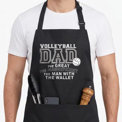 Volleyball english dad apron - Stacked white and black text reading Volleyball Dad The Great The Magnificent The Man — shown on a black cotton canvas apron against a white background