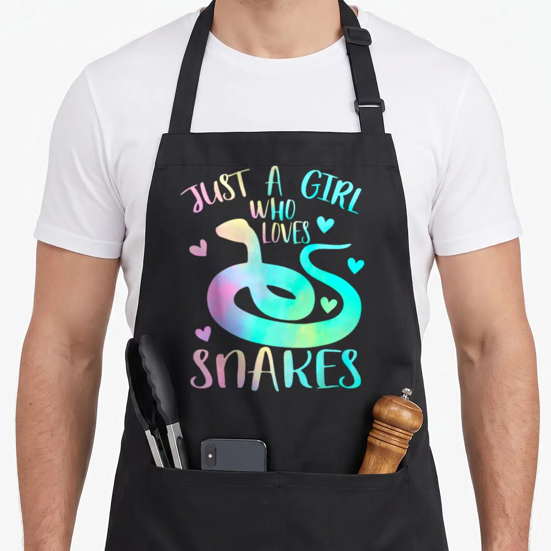 Snake themed aprons - Cartoon snake in pink yellow cyan with four hearts and text JUST A GIRL WHO LOVES SNAKES — shown on a black cotton canvas apron against a white background