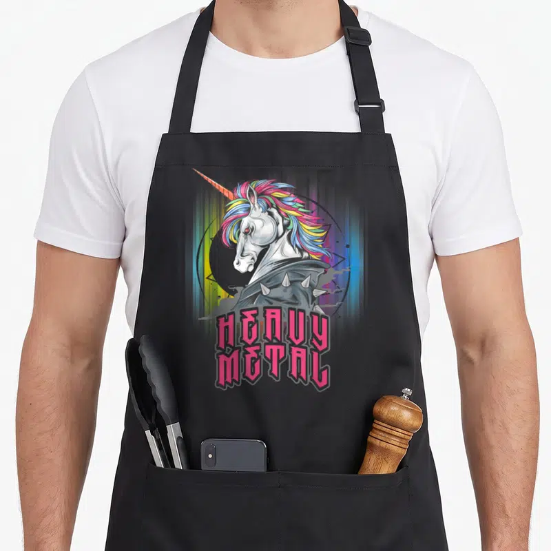 Unicorn heavy apron - Cartoon unicorn with spiked collar and pink heavy metal text — shown on a black cotton canvas apron against a white background