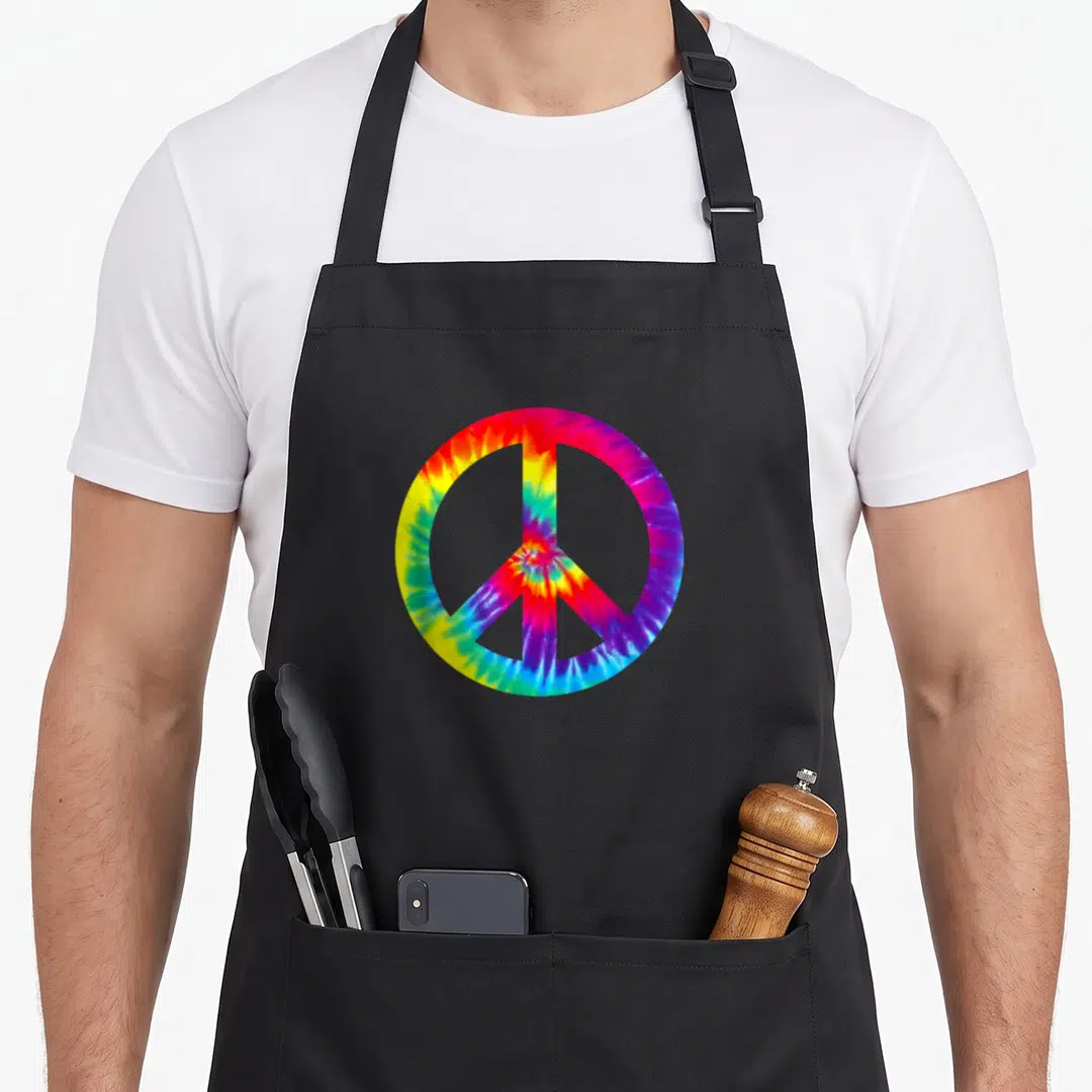 Symbol tie-dye apron - Colorful tie-dye peace symbol print with red yellow green blue purple swirls — shown on a black cotton canvas apron against a white background