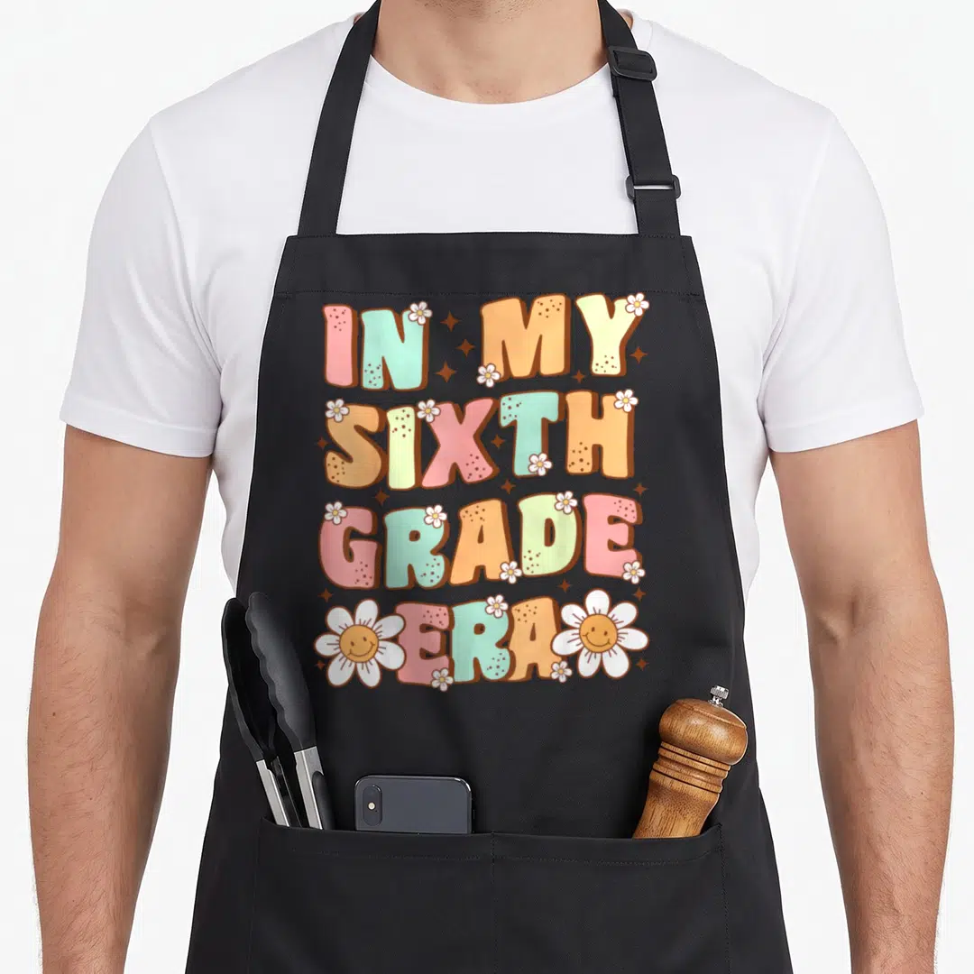 Sixth groovy grade apron - IN MY SIXTH GRADE ERA text in pink orange teal with smiling daisies and stars — shown on a black cotton canvas apron against a white background